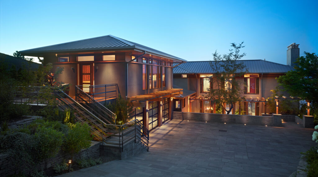 Contemporary stucco and wood house with flat roofs, extensive window walls, metal railings, lit entry stairs and deck overlooking landscaped courtyard with potted plants and driveway.