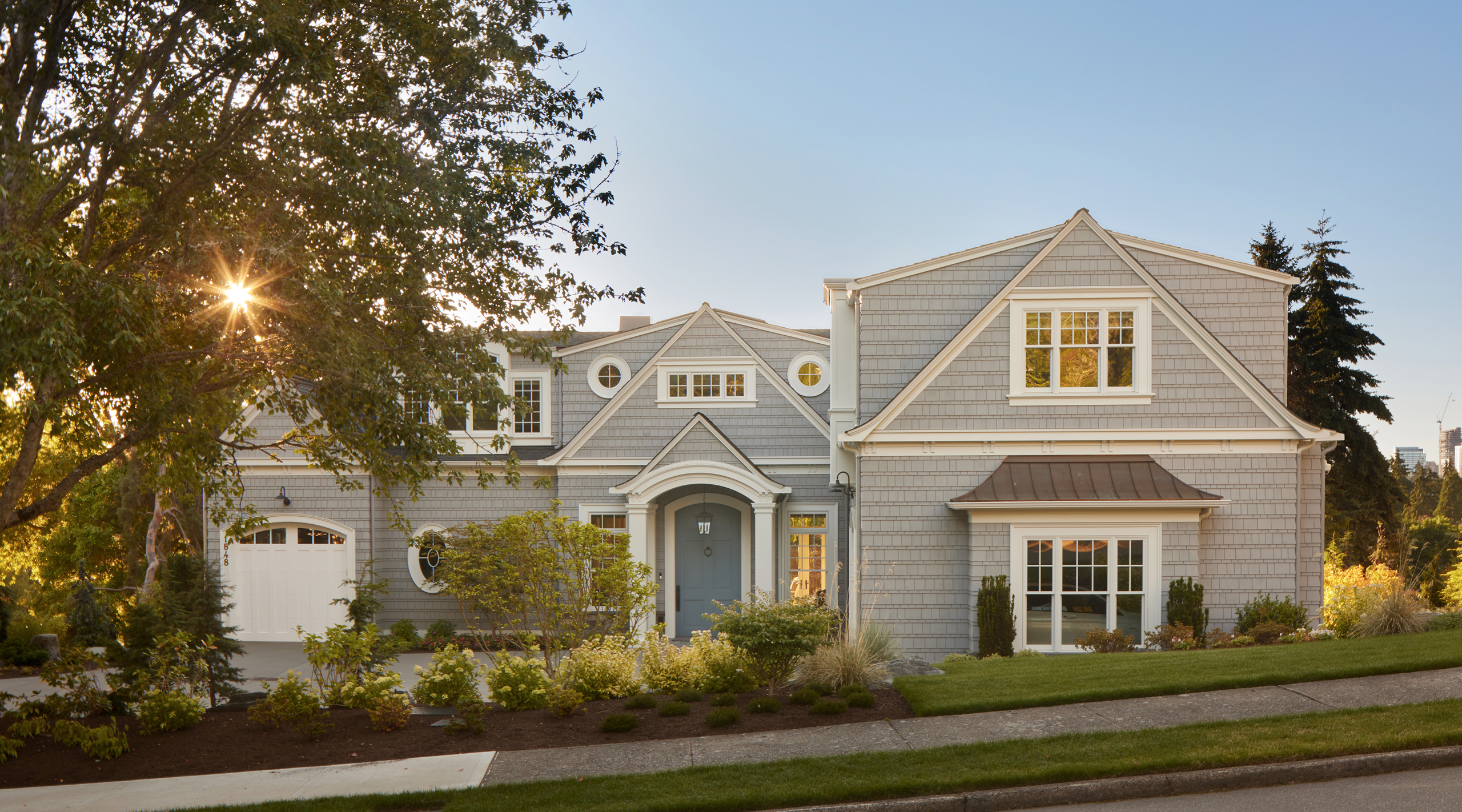 Light gray shingle-style bungalow with gabled dormers and round windows, blue front door under portico, attached garage, driveway lined with grasses and shrubs, trees, setting sun rays.