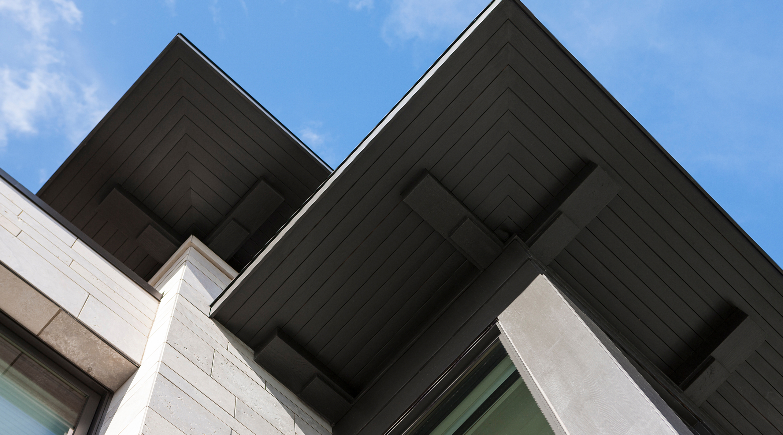 Dramatic architectural detail of intersecting dark metal roofs over white stone building walls and slim vertical windows under expansive blue sky with scattered clouds.